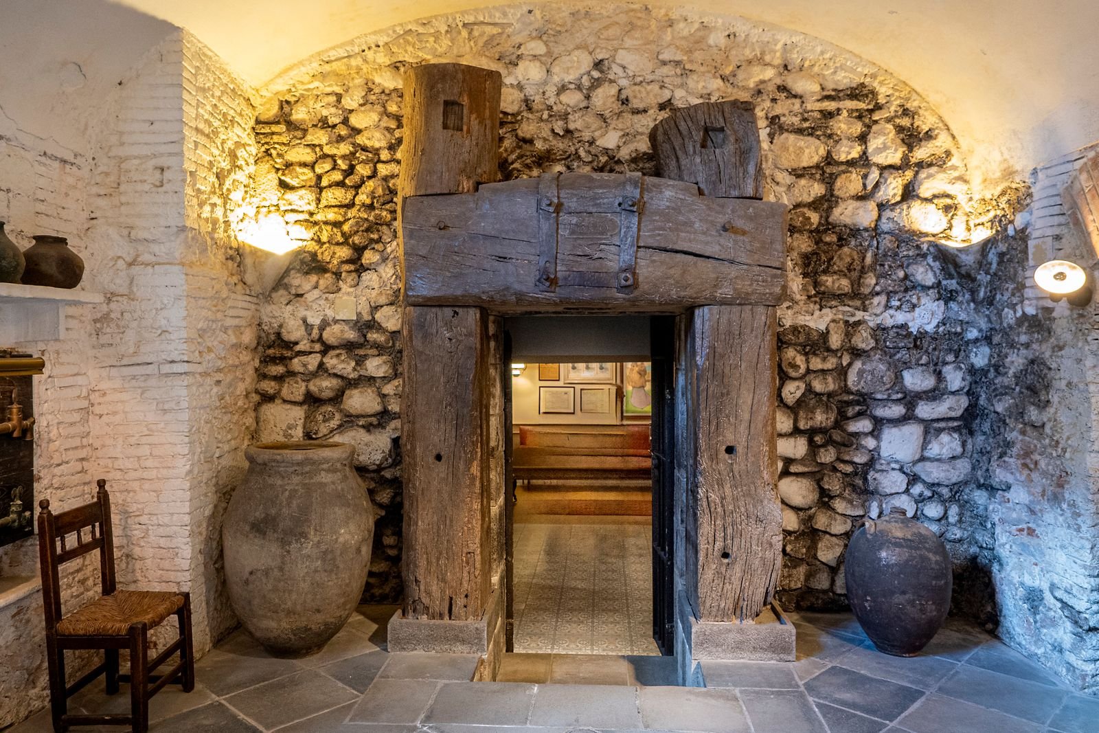 Cave ancienne voûtée en pierre calcaire, éclairée par deux appliques, avec une porte basse encadrée de poutres et un sol dallé irrégulier.