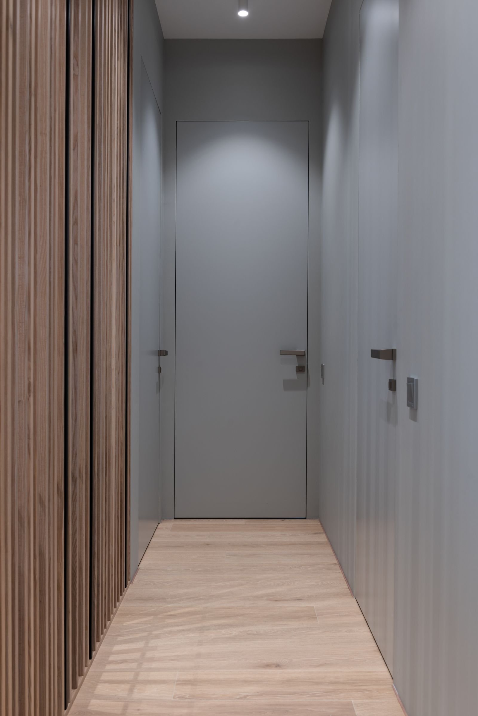 Couloir intérieur étroit et sans fenêtre, bardage bois clair sur un côté, portes affleurantes de l'autre, sol clair et éclairage encastré.
