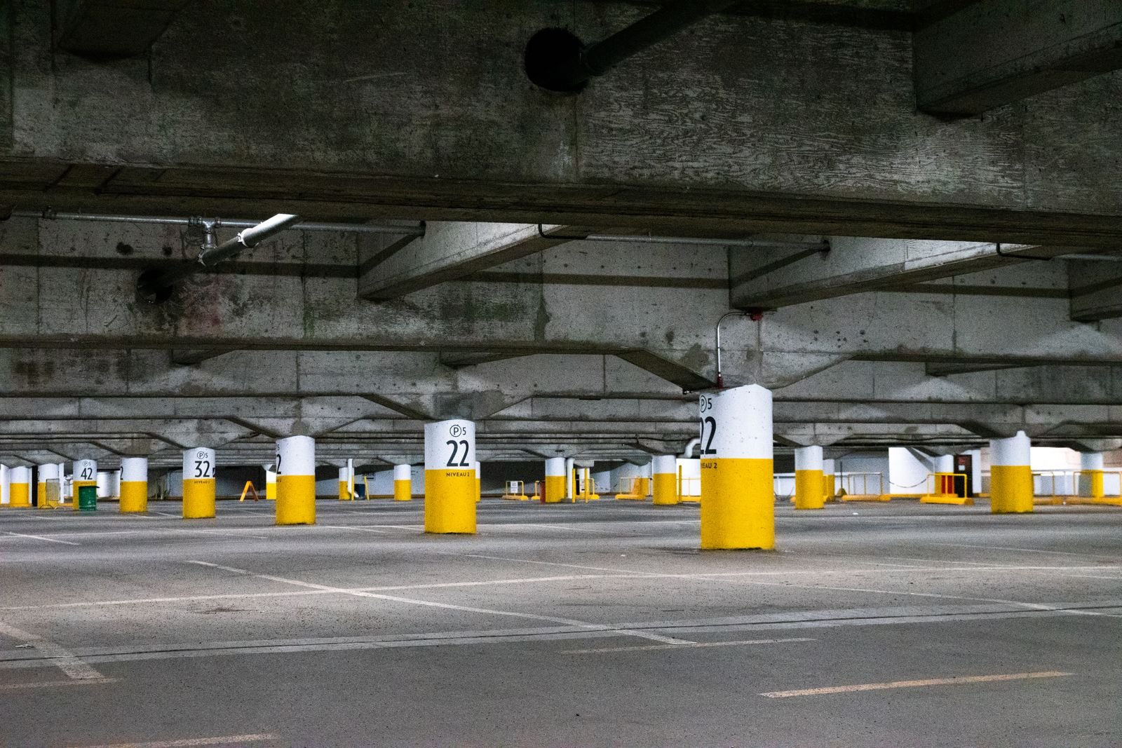 Niveau de parking enterré entièrement vide, piliers de béton à bandes jaunes, poutres apparentes et marquage au sol effacé par endroits.