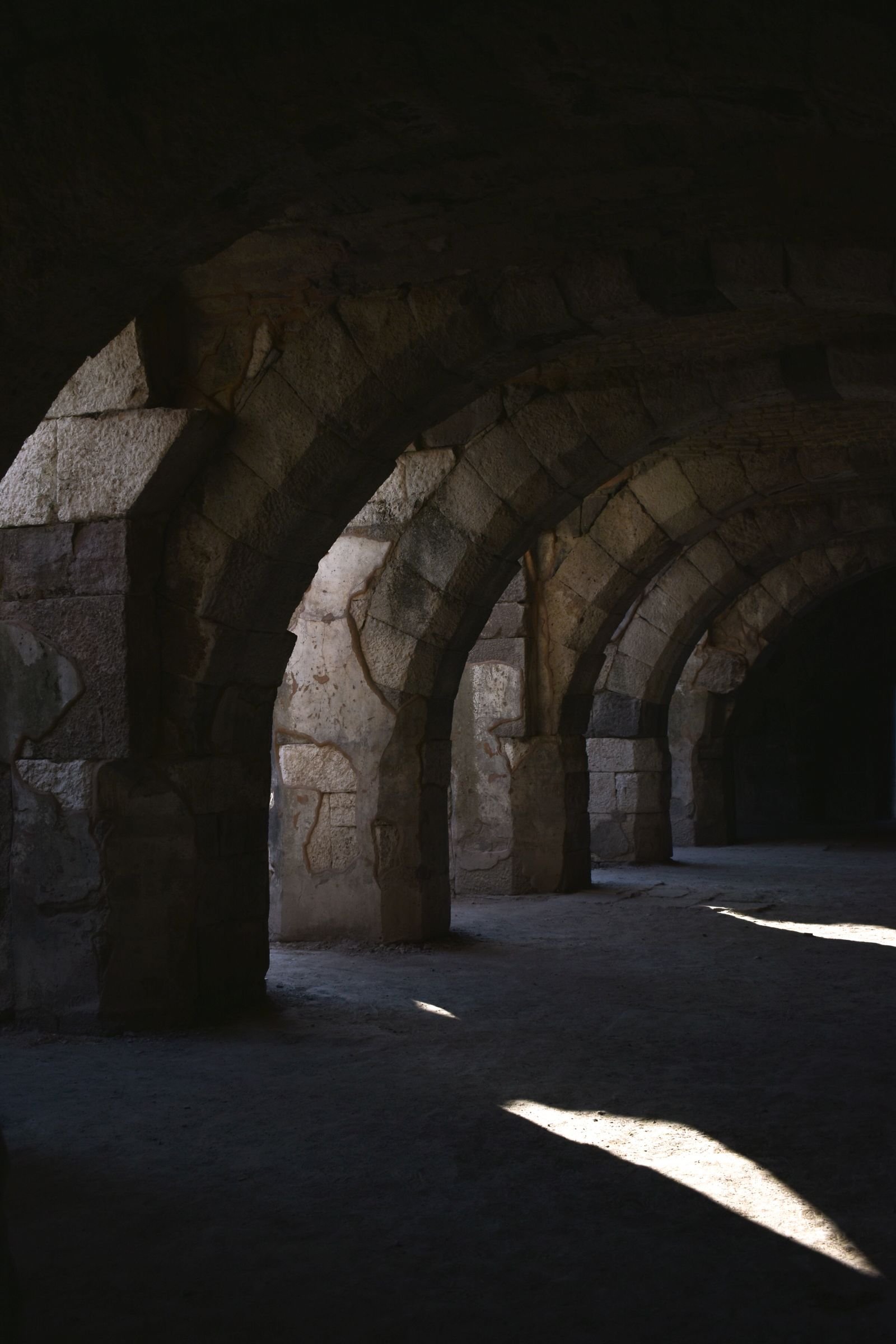 Salle enterrée sombre à voûtes de pierre reposant sur des piliers massifs, seule une tache de lumière naturelle éclaire le sol brut.