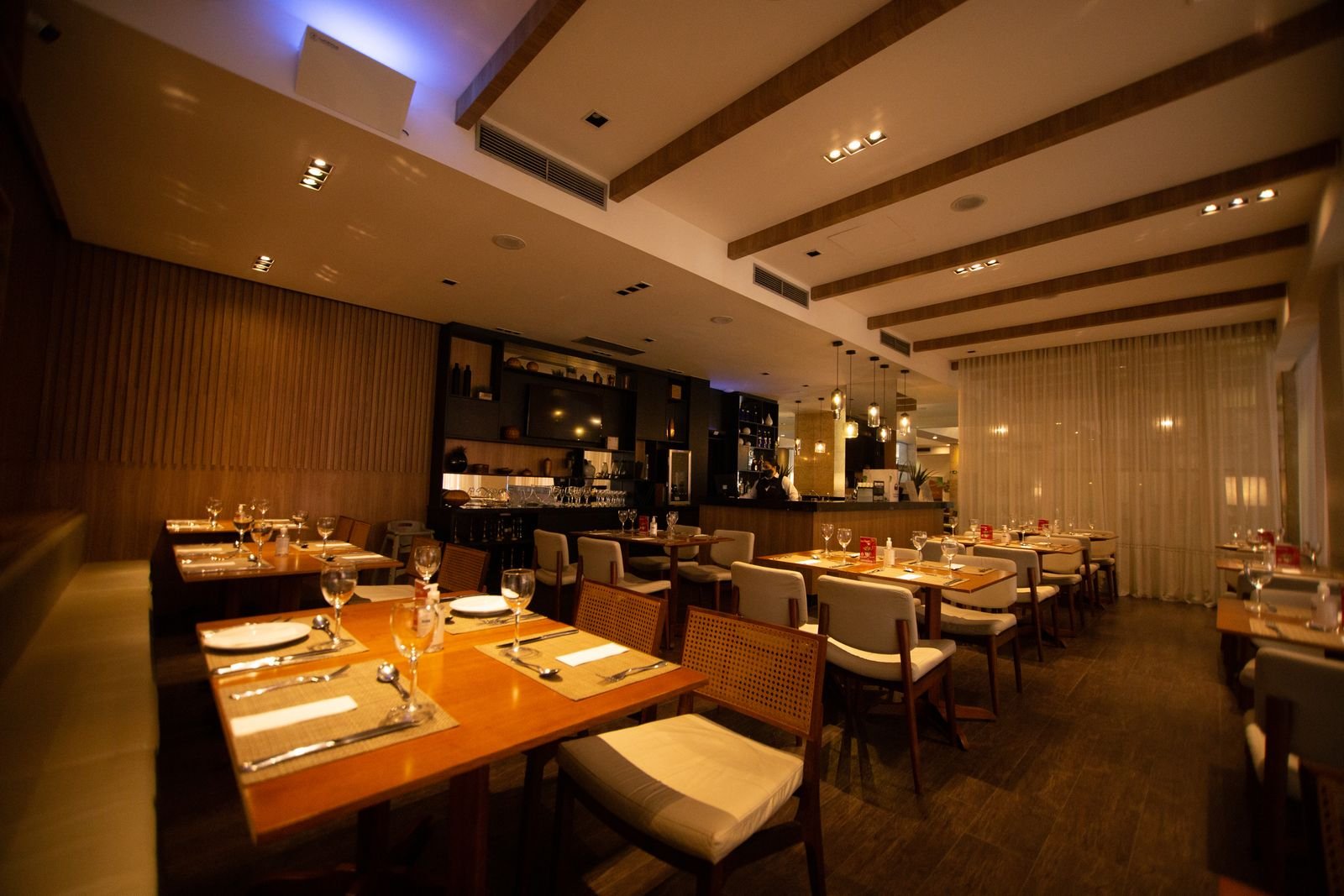 Salle de restaurant vide et entièrement dressée, tables nappées et verres en place, éclairage tamisé et comptoir au fond.